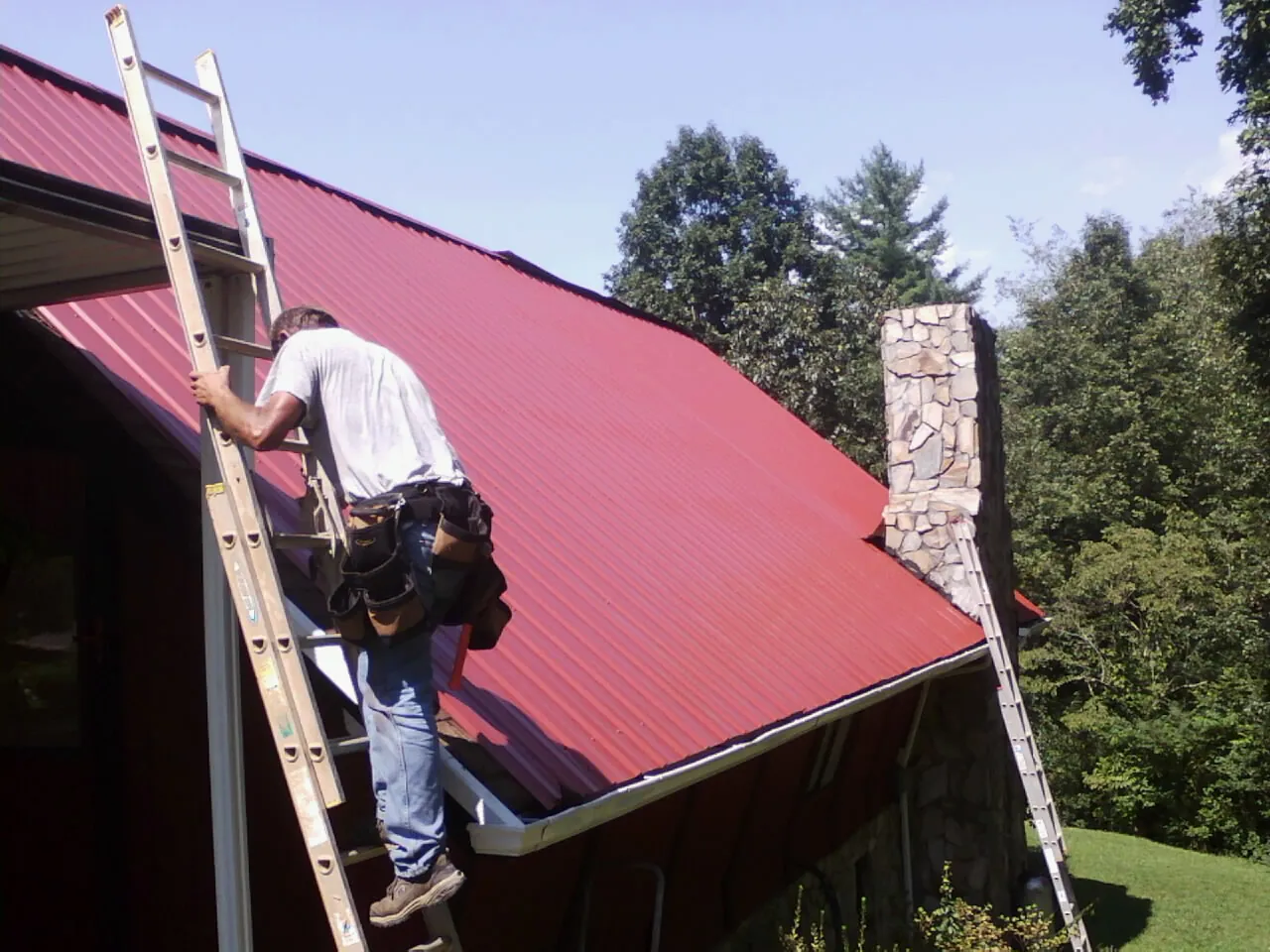 Roofer on a ladder beside a red metal roof and stone chimney — Soffit & Fascia Repair by Roofing Brothers in Greenwood Lake